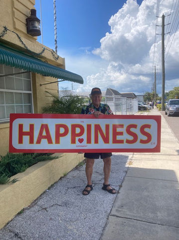 Gary King in a Hawaiian shirt holding a large red HAPPINESS sign on a St. Petersburg sidewalk, with a yellow stucco building and blue sky behind him