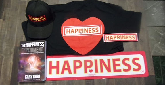 A flat-lay of Happiness Experiment merchandise: the book, a black t-shirt with a red heart and HAPPINESS text, a baseball cap, a small sign, and a full-size red HAPPINESS sign